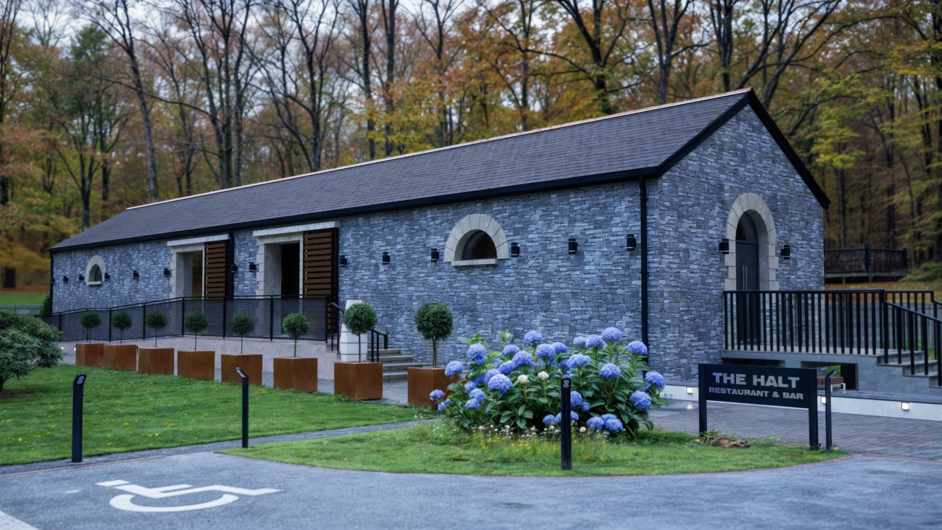 Restaurant & bar building in County Donegal designed by Usonia Architects, housed in a former railway building featuring an original stone facade, arched windows, complemented by a modern extension featuring a curved roof with coloured cladding panels, set amidst lush greenery and tall trees. Outdoor patio area with stylish fencing and planters.