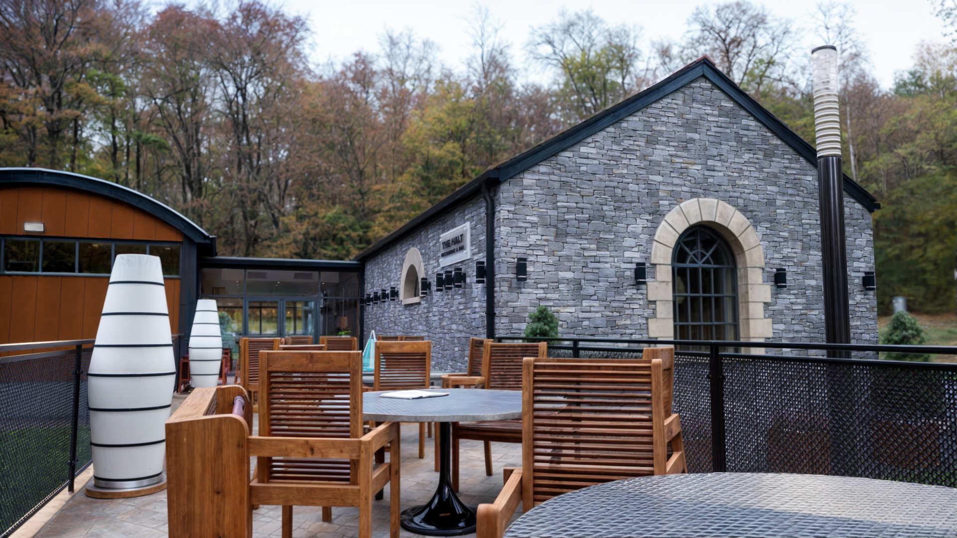 Restaurant & bar building in County Donegal designed by Usonia Architects, housed in a former railway building featuring an original stone facade, arched windows, complemented by a modern extension featuring a curved roof with coloured cladding panels, set amidst lush greenery and tall trees. Outdoor patio area with stylish fencing and planters.