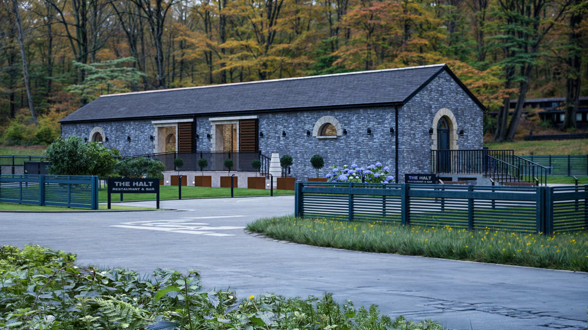 Restaurant & bar building in County Donegal designed by Usonia Architects, housed in a former railway building featuring an original stone facade, arched windows, complemented by a modern extension featuring a curved roof with coloured cladding panels, set amidst lush greenery and tall trees. Outdoor patio area with stylish fencing and planters.