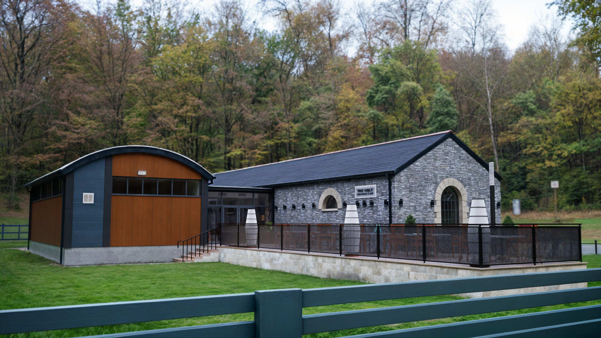 Restaurant & bar building in County Donegal designed by Usonia Architects, housed in a former railway building featuring an original stone facade, arched windows, complemented by a modern extension featuring a curved roof with coloured cladding panels, set amidst lush greenery and tall trees. Outdoor patio area with stylish fencing and planters.